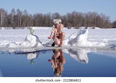 An Adult Cheerful Woman In Faun Costume With Horns On The Edge Of An Ice Hole With Cold Water On Sunny Winter Day. Next To Bouquet Of Snowdrops.