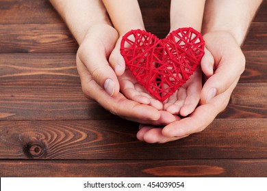 Adult And Child Holding Red Heart In Hands Over A Wooden Table. Happy Family Relationships, Health Care, Pediatric Cardiology Concept.