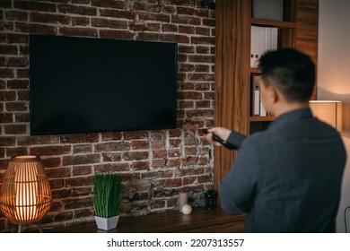 Adult Chinese Man With Remote Control Turns On Tv With Blank Screen In Home Room Interior, Back. Entertainment With Modern Technology, News, Advertising And Good Offer, Movie Or Show At Spare Time