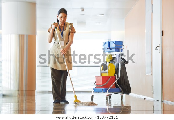 Adult cleaner maid woman\
with mop and uniform cleaning corridor pass or hall floor of\
business building