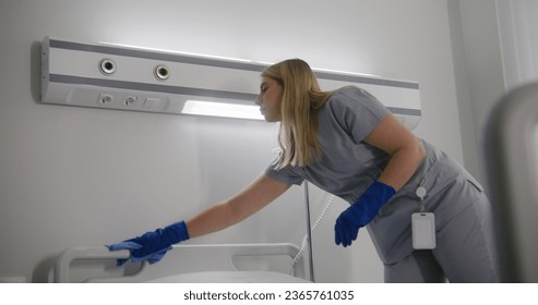 Adult cleaner wipes patient bedside panel and bedside table with rug in hospital ward. Mature health worker changes bedclothes after patients. Nurses clean and prepare hospital room for new patients. - Powered by Shutterstock