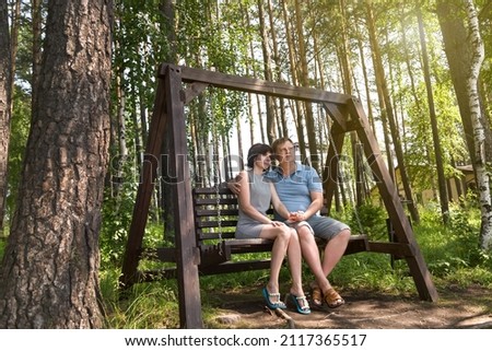 An adult couple in love, a man and a woman, are sitting on a wooden swing in the forest enjoying nature on a summer day.