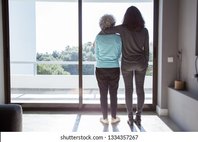 Adult Daughter Hugging Senior Mother. Back View Of Young And Elderly Women Standing And Looking Out Window. Parents Concept