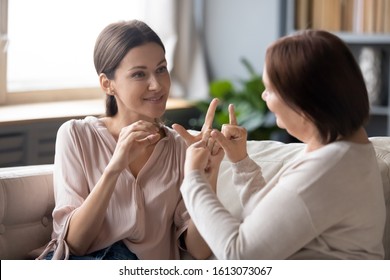 Adult Daughter And Middle-aged Mother Sit On Couch At Home Talk Using Sign Language, Senior Mum Or Teacher Speak Communicate With Grownup Girl Child With Symbols Gestures, Deaf-mute Family Concept