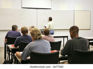 An Adult Education Teacher In Front Of His Class, Drawing A Diagram On The Board.  Focus On Teacher And Diagrams.