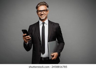 Adult employee IT business man corporate lawyer he wear classic formal black suit shirt tie work in office hold closed laptop pc computer use mobile cell phone isolated on plain grey background studio - Powered by Shutterstock