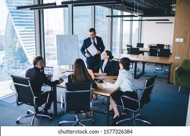 Adult entrepreneur in formal wear standing in conference room and talking with colleagues sitting at table while discussing strategies of company