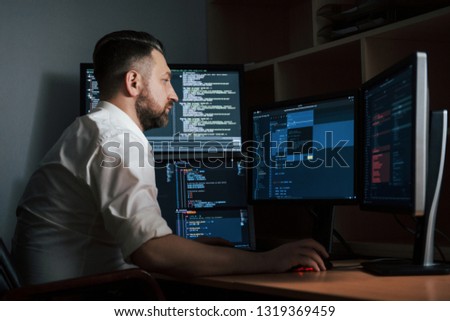 Adult experienced programmer. Bearded man in white shirt works in the office with multiple computer screens in index charts.