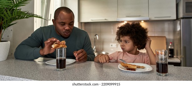 Adult Father And Curly Little Son Having Lunch Or Dinner With Cola And Pizza At Table At Home Kitchen. Unhealthy Eating. Young Black Family Lifestyle And Relationship. Fatherhood And Parenting