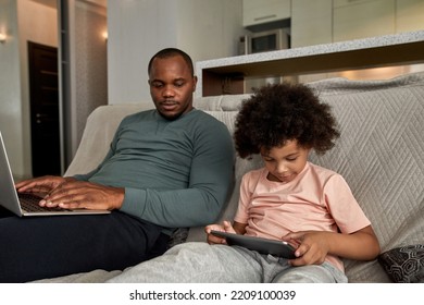 Adult Father With Laptop Looking At His Little Son Using Digital Tablet On Sofa At Home. Young Black Family Lifestyle And Relationship. Domestic Leisure And Entertainment. Fatherhood And Parenting