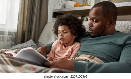 Adult Father And Little Son Reading Book On Sofa Under Blanket At Home. Young Black Family Spending Time Together. Fatherhood And Parenting. Domestic Leisure, Hobby And Entertainment