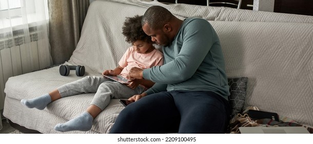 Adult Father And Little Son Using Digital Tablet On Sofa At Modern Home. Young Black Family Lifestyle And Relationship. Domestic Leisure And Entertainment. Fatherhood And Parenting