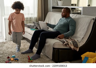 Adult Father Looking At His Offended Curly Little Son Going On Carpet At Home. Man Working On Laptop Computer On Sofa. Young Black Family Lifestyle And Relationship. Fatherhood And Parenting