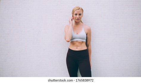 Adult Female Athlete In Sports Bra And Leggings Touching Hair While Standing Next To Light Grey Wall And Looking At Camera