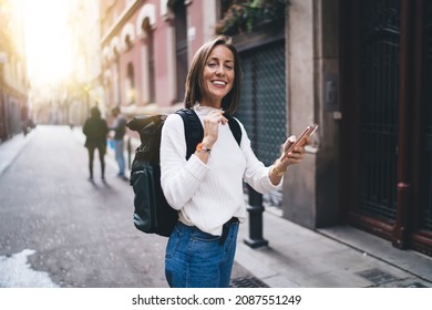 Adult female with brown hair in casual clothes with backpack browsing phone while wandering streets of city and looking at camera - Shutterstock ID 2087551249