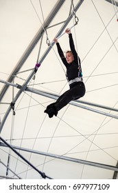 An adult female hangs on a flying trapeze at an indoor gym. The woman is an amateur trapeze artist. 