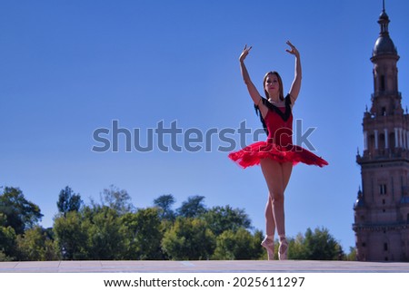 Adult female Hispanic classical ballet dancer in red tutu doing figures in the middle of a plaza on a sunny day.