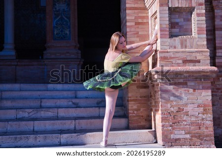 Adult female Hispanic classical ballet dancer in green and black tutu with coins, performing figures beside steps next to a brick pillar.