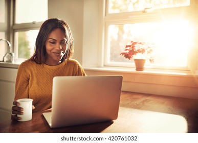 Adult Female Holding Cup And Laughing While Looking At Her Laptop Computer While Sitting Near Bright Sunny Window In Kitchen