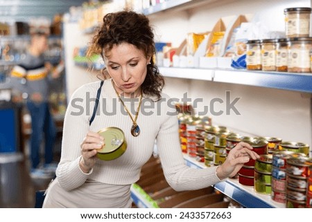 Adult female shopper in casual clothes chooses canned sprats in grocery store