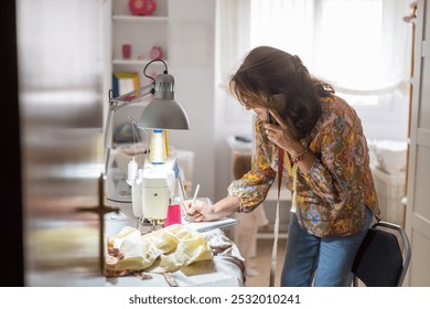 Adult Female Tailor Working from Home with Grandchild, Managing Orders in Workshop.Seamstress at Home Office Taking Orders, Caring for Grandchild.Home-Based Fashion Designer Balancing Work and Family. - Powered by Shutterstock