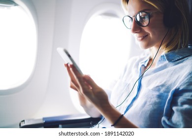 Adult female using phone with earphones while sitting on plane in comfortable chair near window and wearing glasses and blue shirt