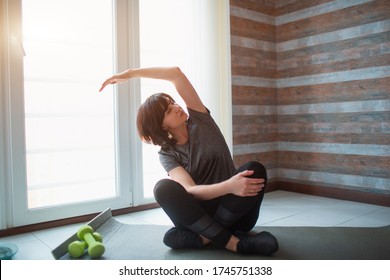 Adult Fit Slim Woman Has Workout At Home. Senior Model Sit With Legs Crossed And Stretches Arm To Side. Exercising Alone To Get In Good Shape And Better Health.