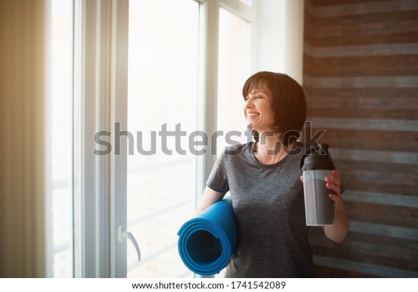 Adult fit slim woman has workout at\
home. Stand after training with some protein drink and yoga mat in\
hands. Care about slim good body shape and well\
being.