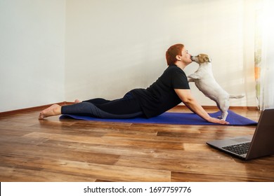 Adult girl with a dog Jack Russell practice online yoga lesson at home during quarantine isolation during the coronavirus pandemic. Dog licks the girl face, flirts.