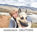 Adult girl with shepherd dog taking selfie near water of lake or river in mountain. Middle aged woman and big pet on nature. Friendship, love and fun