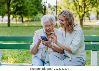 Adult granddaguhter helping her grandmother to use cellphone when sitting on bench in park in summer. - Shutterstock ID 2172757279