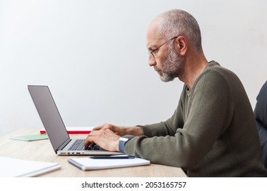 Adult Gray-haired Man Working On A Laptop At A Desk, Photo In Profile