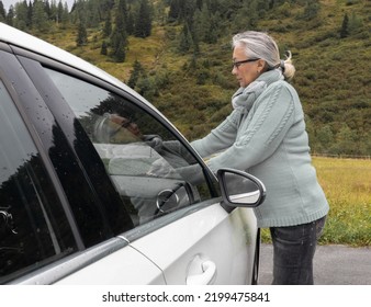 An Adult Gray-haired Woman In A Warm Sweater And Glasses Adjusts The Windshield Wipers Of A Car