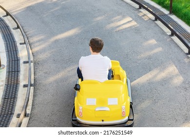 An Adult Guy Is Driving A Small Yellow Children's Car Along A Park Road. Family Holiday. Ride On Cars With The Kids