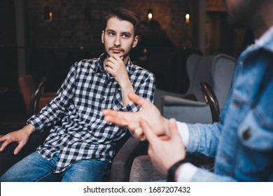 Adult Handsome Male In Casual Outfit Sitting On Chair Touching Chin While Listening To Man In Conversation Having Meeting In Modern Cafe 