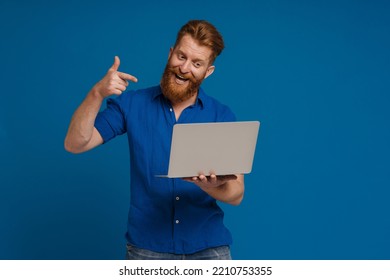 Adult Handsome Redhead Bearded Enthusiastic Man With Open Mouth Holding Laptop, Looking And Pointing At It While Standing Over Isolated Blue Background