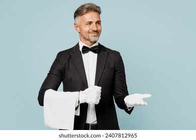 Adult happy barista male waiter butler man wear shirt black suit bow tie gloves elegant uniform hold towel point hand aside work at cafe isolated on plain blue background. Restaurant employee concept