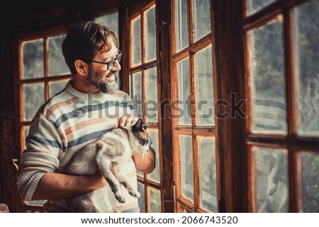 Adult happy mature man with a cat look outside the windows at home enjoying indoor leisure relax and activity alone. Hipster male people with window light smiling in living room