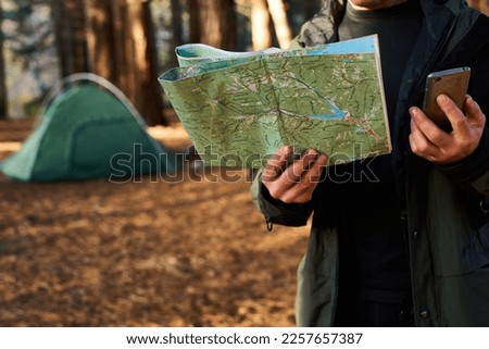 Adult hiker with backpack using mobile phone and map in forest near his tent. Hiking elderly man in autumn nature holding map outdoors next to a campsite in the sunlight closeup shot. Ageless travel