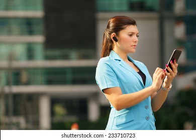 Adult Hispanic Person With Mobile Phone And Wireless Bluetooth Headset, Talking On Telephone In The Street Out Office Buildings 