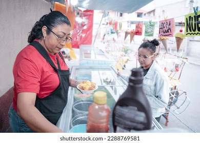 An adult Hispanic woman is preparing a vanilla ice cream for a young woman client in a street market stall