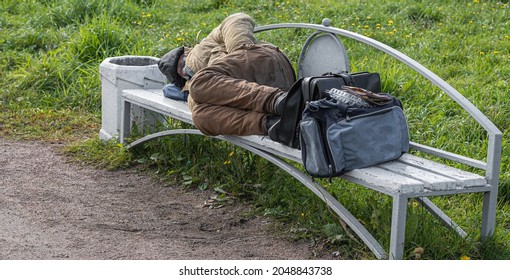 an adult homeless tramp sleeps on a park bench in the open air next to a bag and briefcase. - Shutterstock ID 2048843738