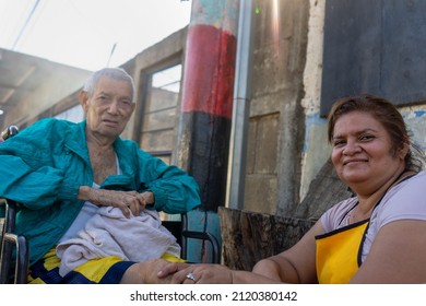 Adult Latin Woman With Her Elderly Father In A Wheelchair On The Outskirts Of A Poor Neighborhood In Latin America
