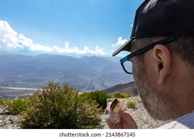 Adult Male (45-50 Years Old) In Profile Looking At The Mountains, With Black Cap And Glasses, And Snack In Hand (alfajor, Biscuit)..