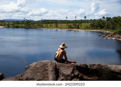 adult male backwards shirtless with hat sitting on edge of rock in Salto El Sapo waterfall with beautiful view of Canaima lagoon, Carrao river, Canaima National Park, Venezuela - Powered by Shutterstock