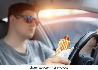 Adult Male Caucasian Person Holding Hot Dog Fast Food Snack While Driving Car On Sunny Day. Men Eating Food While Driving Steering Wheel. Distracted Man In Vehicle. Bad Unhealthy Habit. Junk Food Go