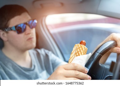 Adult Male Caucasian Person Holding Hot Dog Fast Food Snack While Driving Car On Sunny Day. Men Eating Food While Driving Steering Wheel. Distracted Man In Vehicle. Bad Unhealthy Habit. Junk Food Go