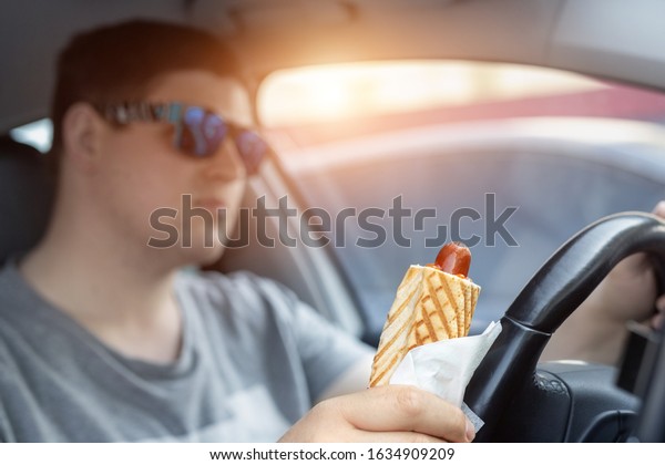 Adult male caucasian person holding hot dog fast food\
snack while driving car on sunny day. Men eating food while driving\
steering wheel. Distracted man in vehicle. Bad unhealthy habit.\
Junk food go