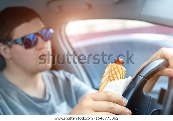 Adult male caucasian person holding hot dog fast food\
snack while driving car on sunny day. Men eating food while driving\
steering wheel. Distracted man in vehicle. Bad unhealthy habit.\
Junk food go