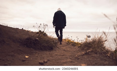 An adult male with a hoodie walking along a sandy path by the shore on a cloudy day - Powered by Shutterstock